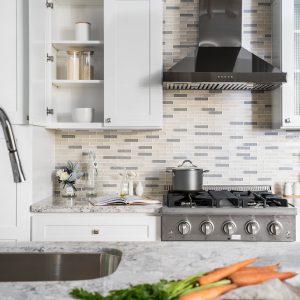 White shaker kitchen cabinets with gray and white geometric mosaic tile backsplash, stainless steel black hood vent, gas cooktop, and gray marble countertop with fresh vegetables