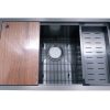 Stainless steel kitchen sink with wooden cutting board, metal grid, and black strainer accessories