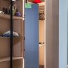 Modern wooden closet interior with glass door, illuminated shelves displaying accessories, clothing on hangers, red cap, and blue panel mirror