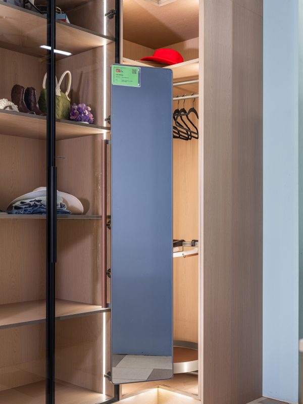 Modern wooden closet interior with glass door, illuminated shelves displaying accessories, clothing on hangers, red cap, and blue panel mirror