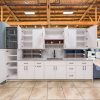 White kitchen cabinet island with gray quartz countertop, open shelving, glass-front refrigerator, and multiple cabinet doors in warehouse showroom