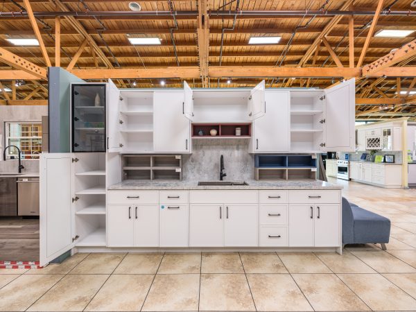 White kitchen cabinet island with gray quartz countertop, open shelving, glass-front refrigerator, and multiple cabinet doors in warehouse showroom