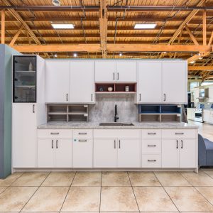 White kitchen cabinet display with closed cabinet doors, gray countertop, open shelf units, and integrated sink in warehouse setting