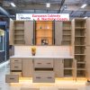 Light wood European cabinet with open door displaying interior shelving, white countertop, glass-front center cabinet, and yellow base lighting in showroom