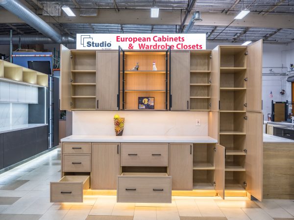 Light wood European cabinet with open door displaying interior shelving, white countertop, glass-front center cabinet, and yellow base lighting in showroom