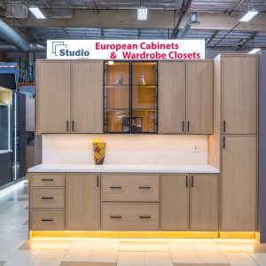 Modern light wood European cabinet display with white countertop, glass-front upper cabinets, closed lower cabinets, and yellow accent lighting in showroom