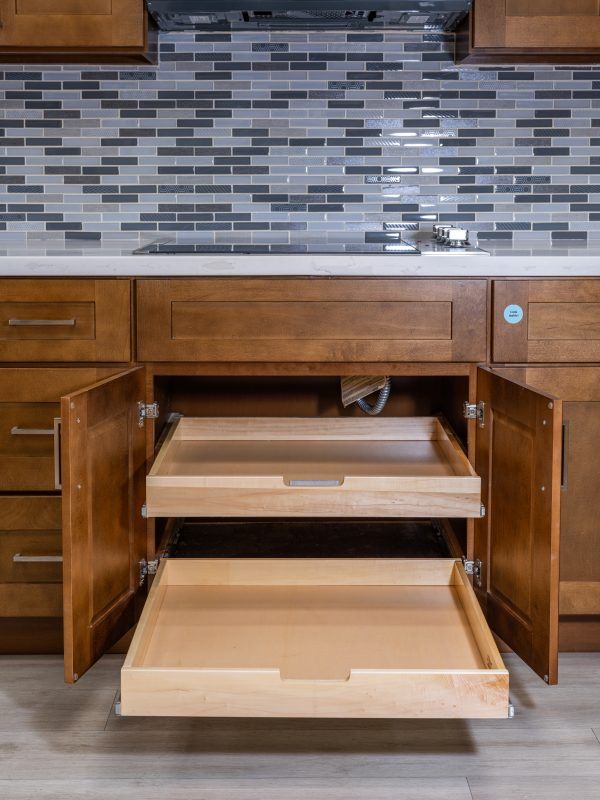 Kitchen cabinet with two wooden pull-out drawers partially extended, displaying natural wood interior construction and metal hardware against gray tile backsplash