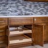 Close-up of open wooden kitchen cabinet with two pull-out drawers and gray tile backsplash above