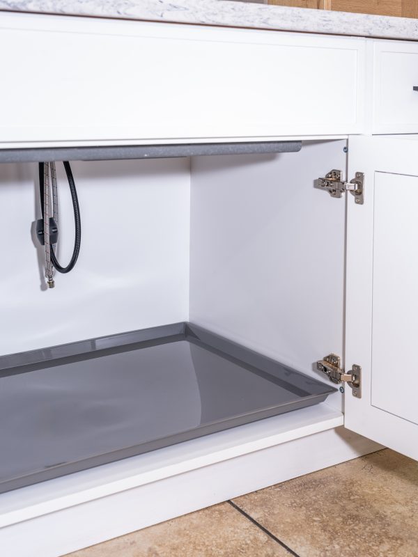 Open white kitchen cabinet with gray pull-out tray and stainless steel hinges, showing interior storage space and coiled black hose