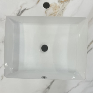 White rectangular ceramic undermount sink with overflow hole and faucet mounting hole, viewed from above on marble surface